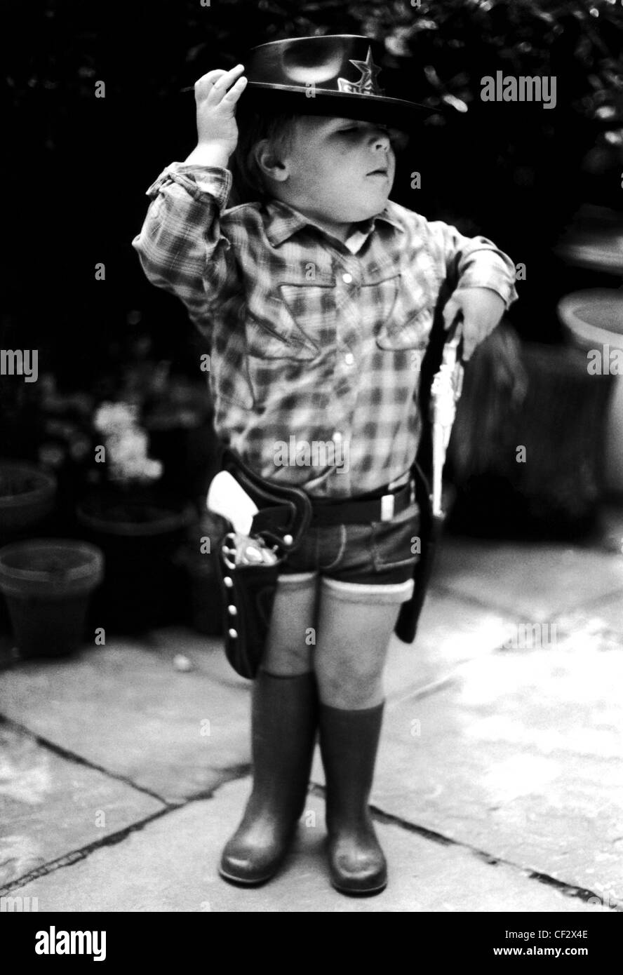 S bambino maschio indossa controllata T-shirt, jeans corti, gomma stivali da pioggia, cinghia fondine pistola e pistole giocattolo e il nero Foto Stock