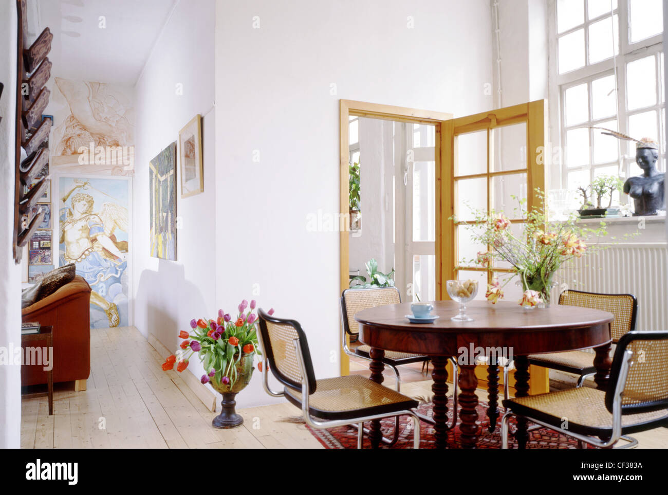 La zona pranzo con tavolo e sedie, un grande vaso di tulipani sul pavimento, quadri sui muri e grandi finestre e legno flowith rug Foto Stock