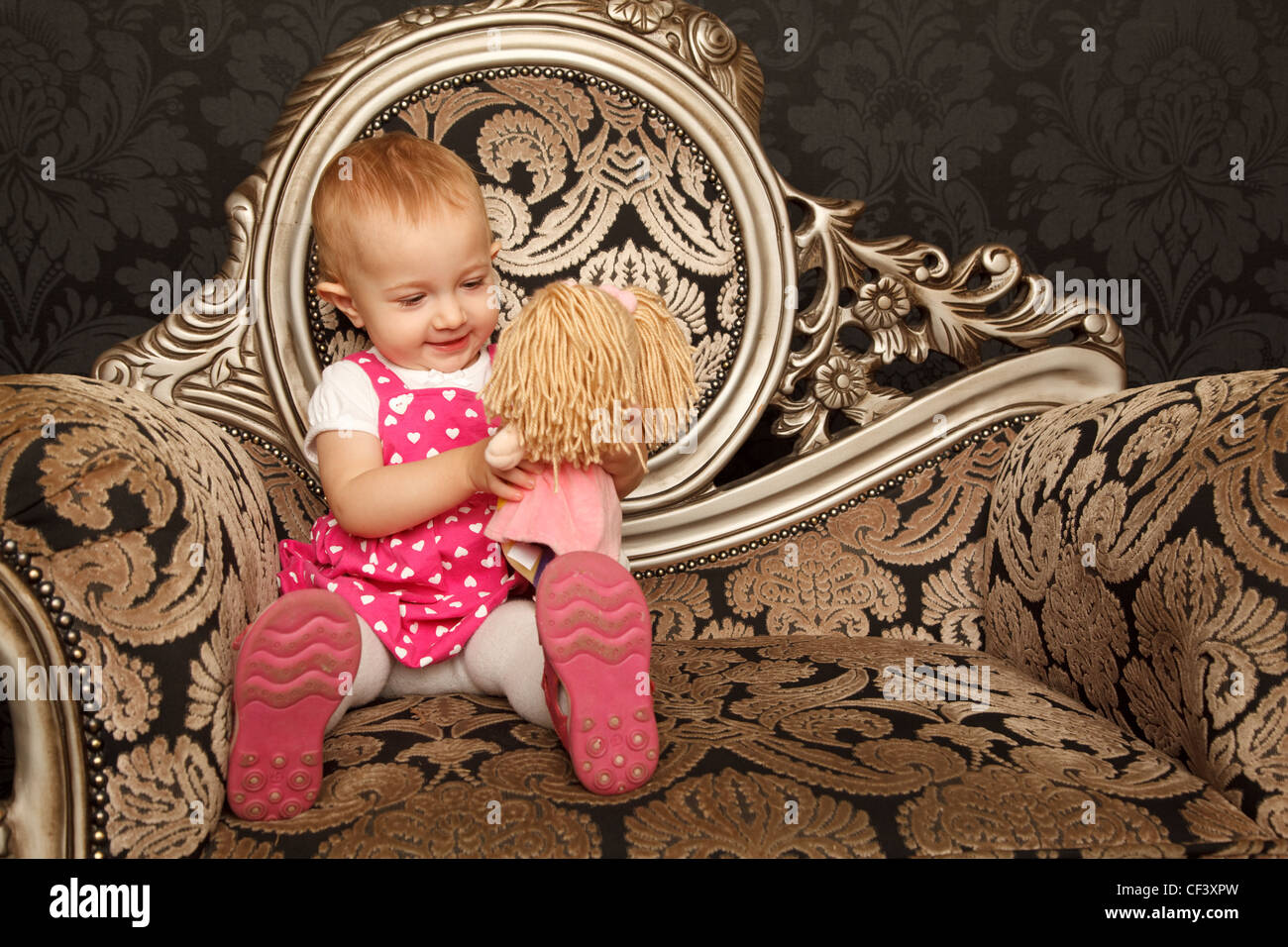 Bambina in abito rosso seduta sulla poltrona retrò con bambola in mani. Formato orizzontale. Foto Stock