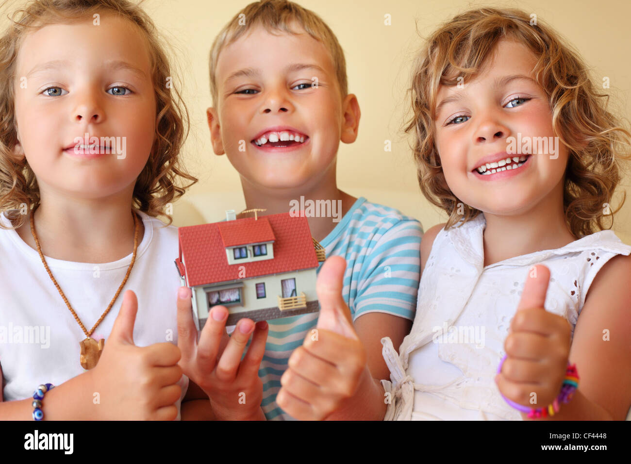 Bambini tre insieme tenendo le mani modello di casa in camera accogliente, mostra ок gesto Foto Stock
