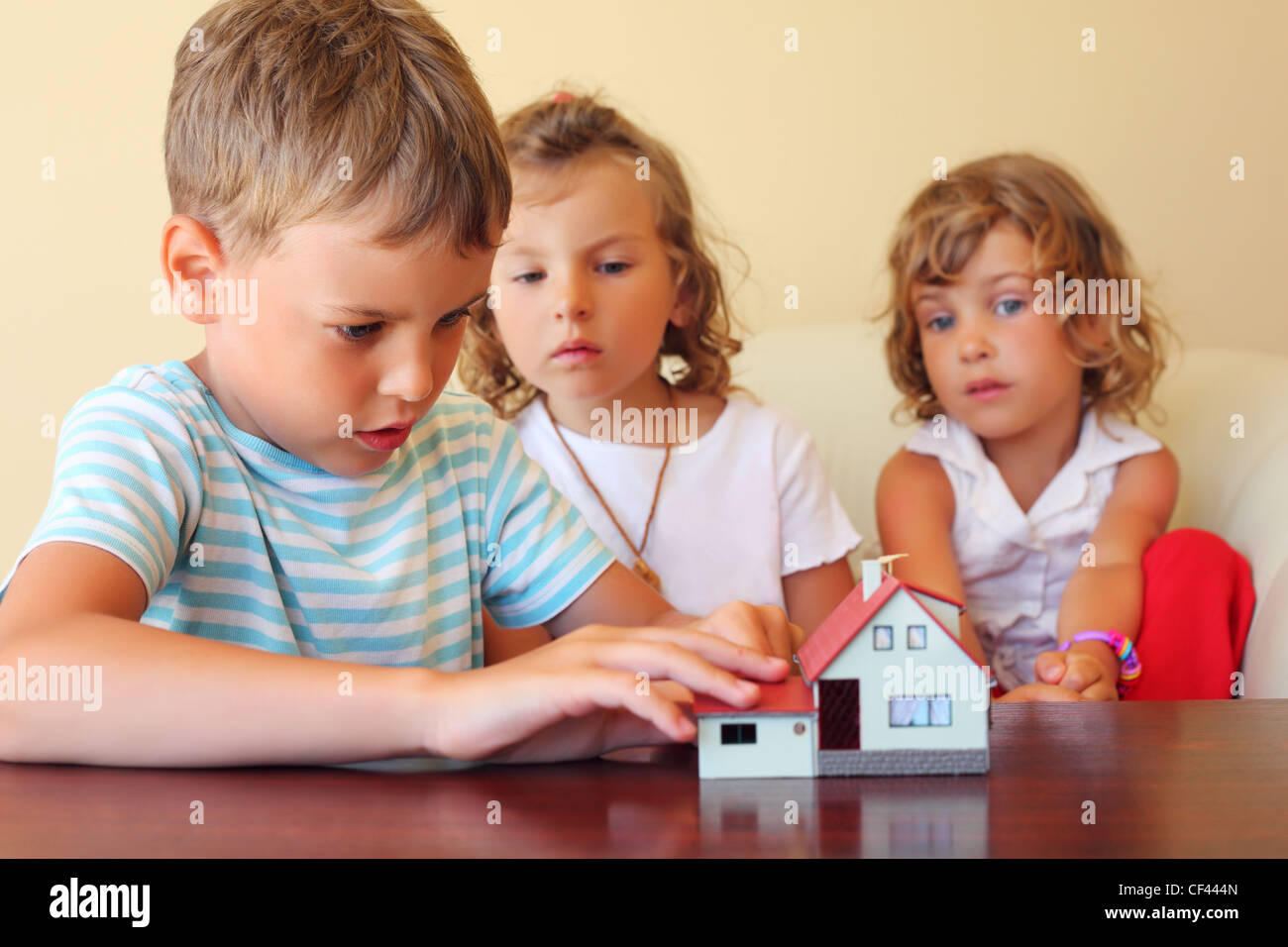 Bambini tre insieme guardando al modello di casa in piedi sulla tavola in camera accogliente Foto Stock