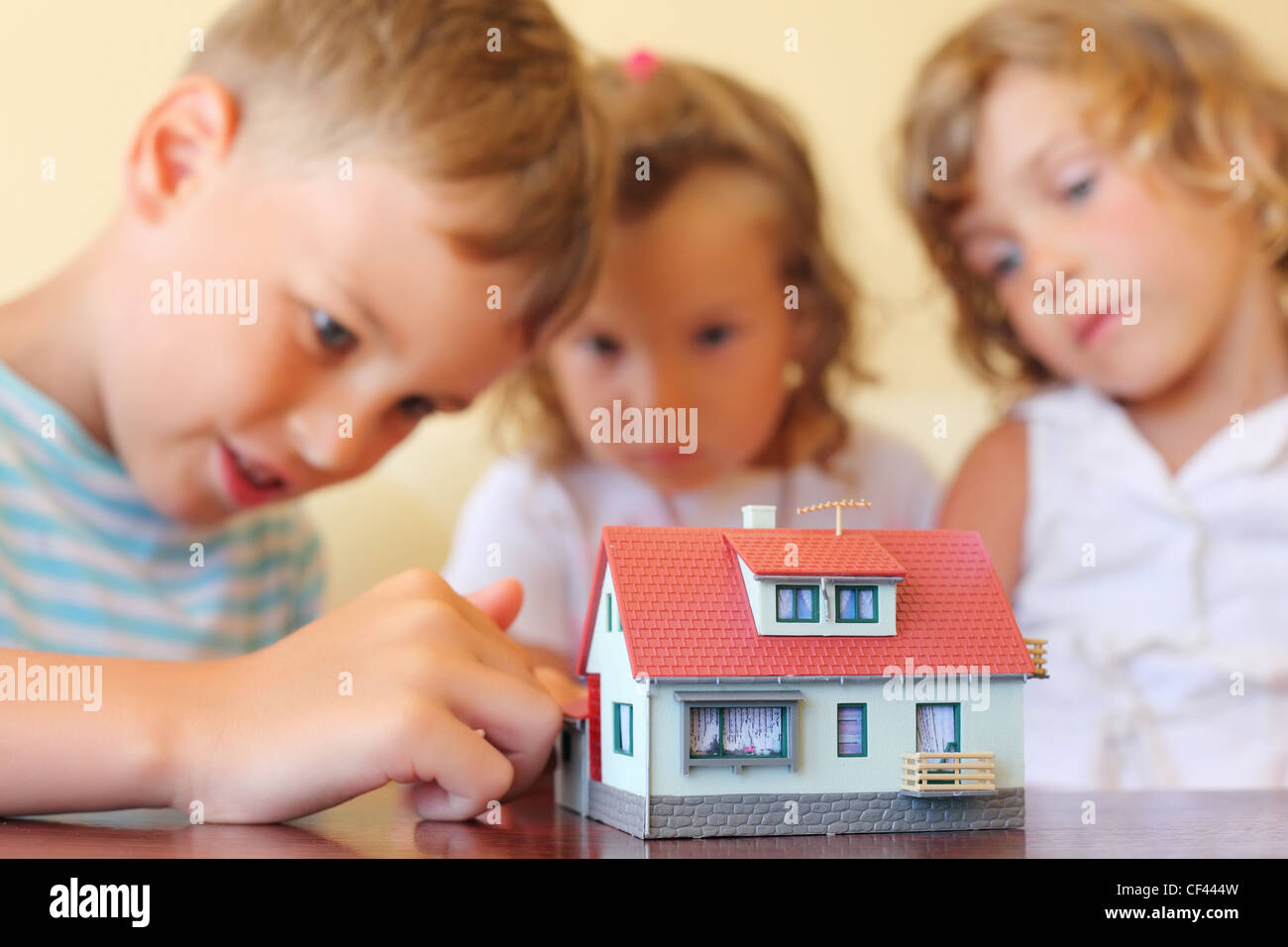 Bambini tre insieme guardando al modello di casa in piedi sulla tavola in camera accogliente, focus sulla casa Foto Stock