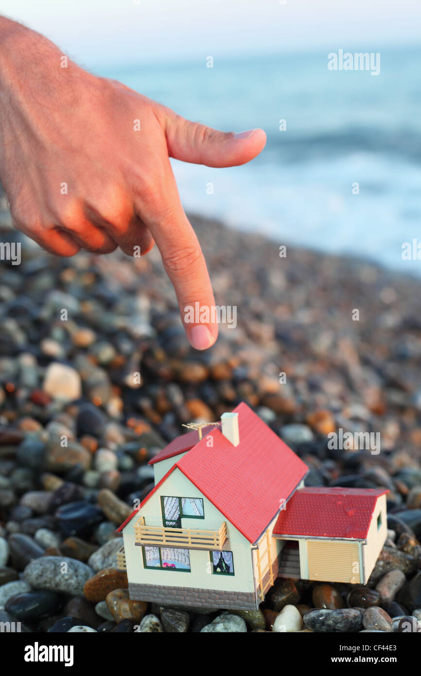 Modello di casa con garage sulla spiaggia sassosa in sera, l'uomo la mano con il dito indice sulla casa Foto Stock