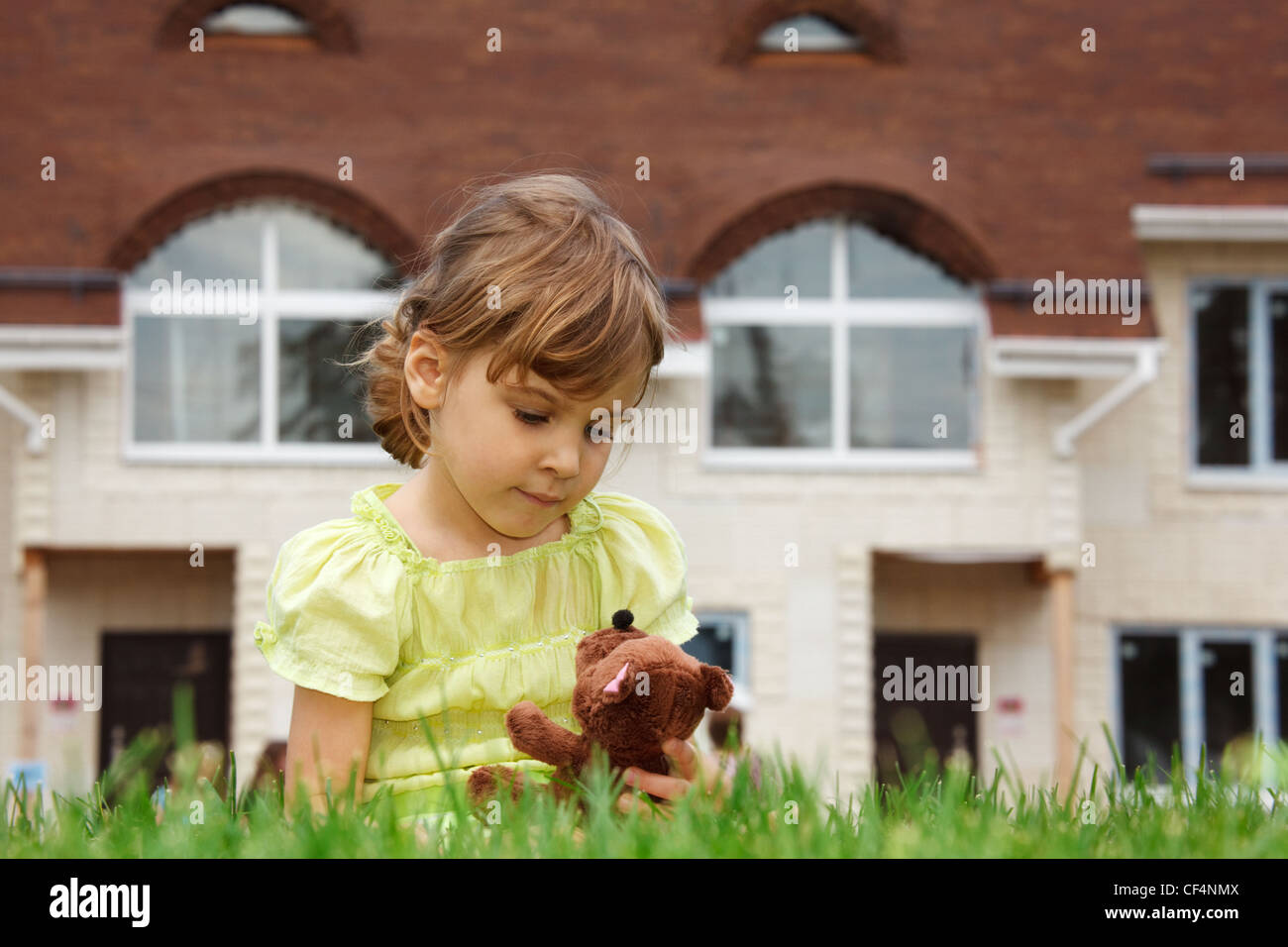 Poco ragazza seduta sul prato davanti la casa nuova. Nelle sue mani detiene il giocattolo orsacchiotto di peluche. Foto Stock
