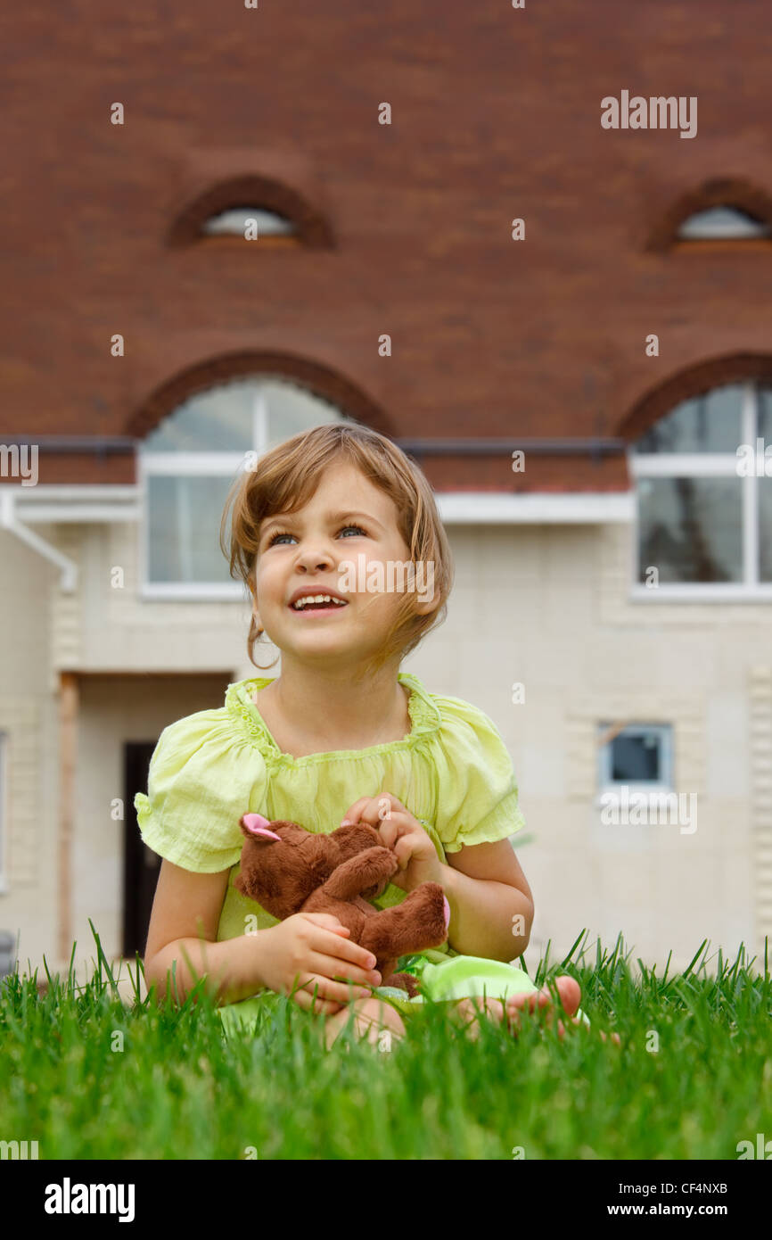 Poco ragazza seduta sul prato davanti la casa nuova. Nelle sue mani detiene toy wolf. formato verticale Foto Stock