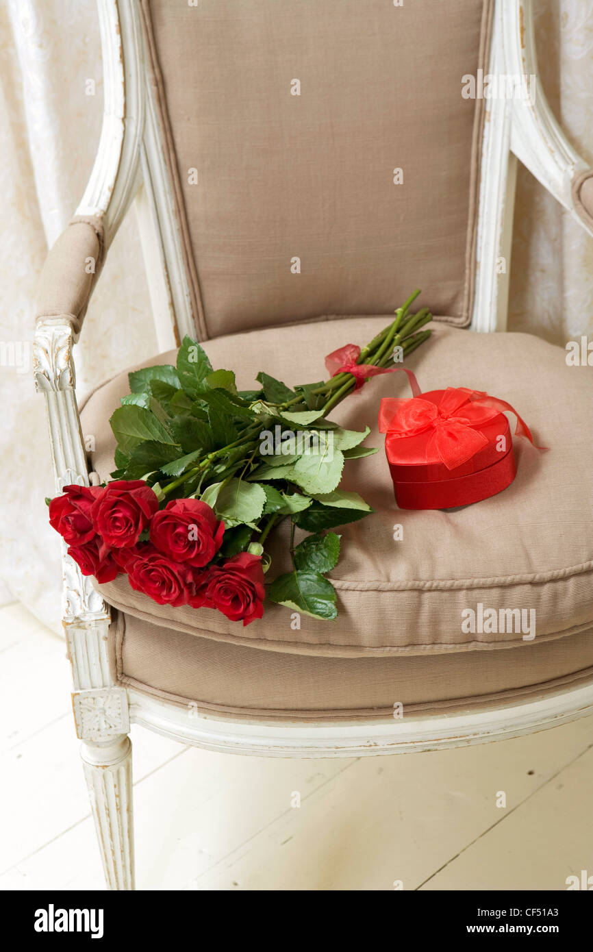 Bouquet di rosso con gambo lungo rose e un cuore rosso a forma di nastro di casella di posta sul sedile di un bianco Edwardian sedia beige Foto Stock