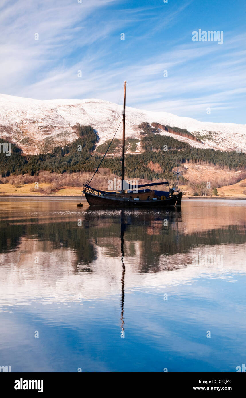 Una barca a vela e delle montagne innevate riflette in Loch Leven, Glencoe. Foto Stock