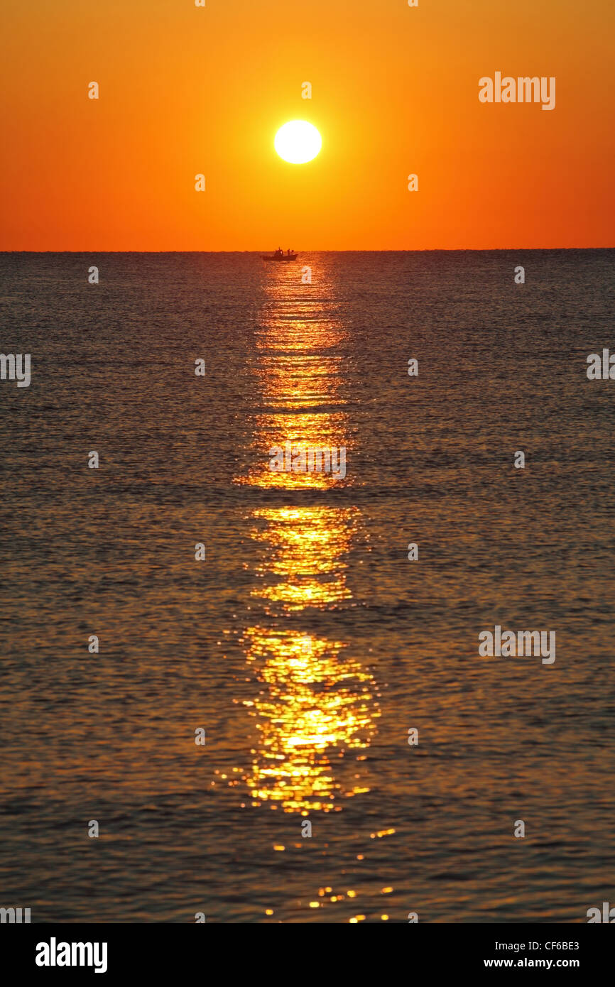 Sun si siede per la linea di orizzonte in mare e si illumina di piccola barca Foto Stock