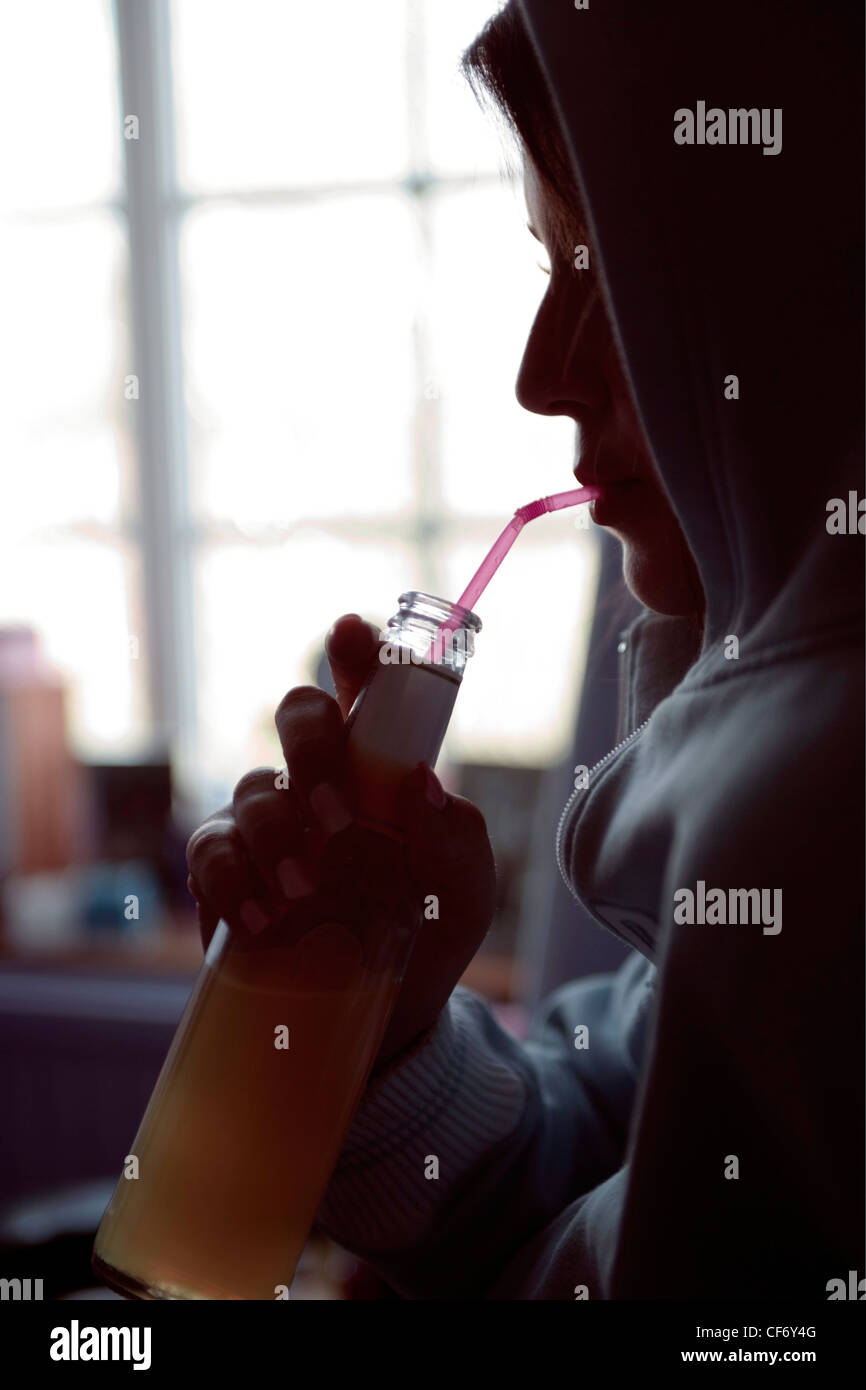 Profilo di Silhouette di adolescente di sesso femminile di indossare top con cappuccio di bere attraverso la paglia dal flacone di alcopop, finestra in background Foto Stock