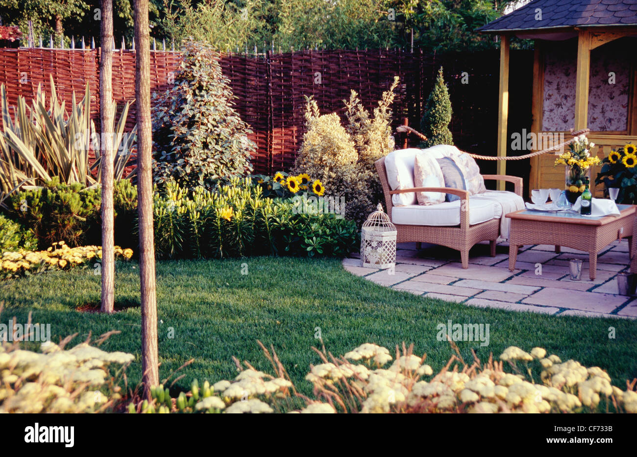 Piccolo giardino ben curato con aiuole di fiori, pavimentato area salotto all pagoda, vimini mobili da giardino, dei bicchieri e delle bottiglie sul tavolo Foto Stock