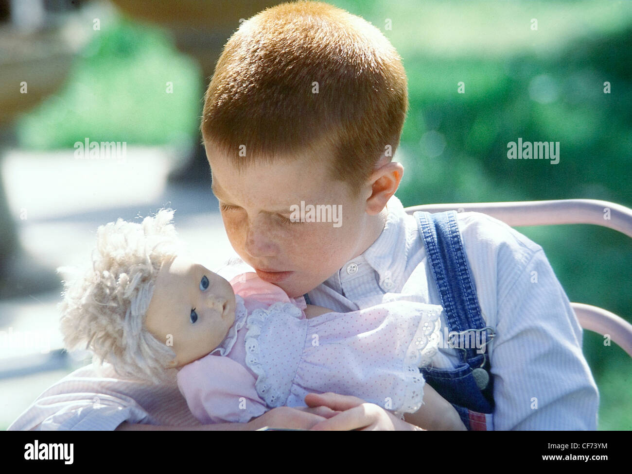 Bambino maschio con breve capelli allo zenzero, indossando un blu pallido camicia a righe e denim complessivamente, abbracciando una bambola Foto Stock
