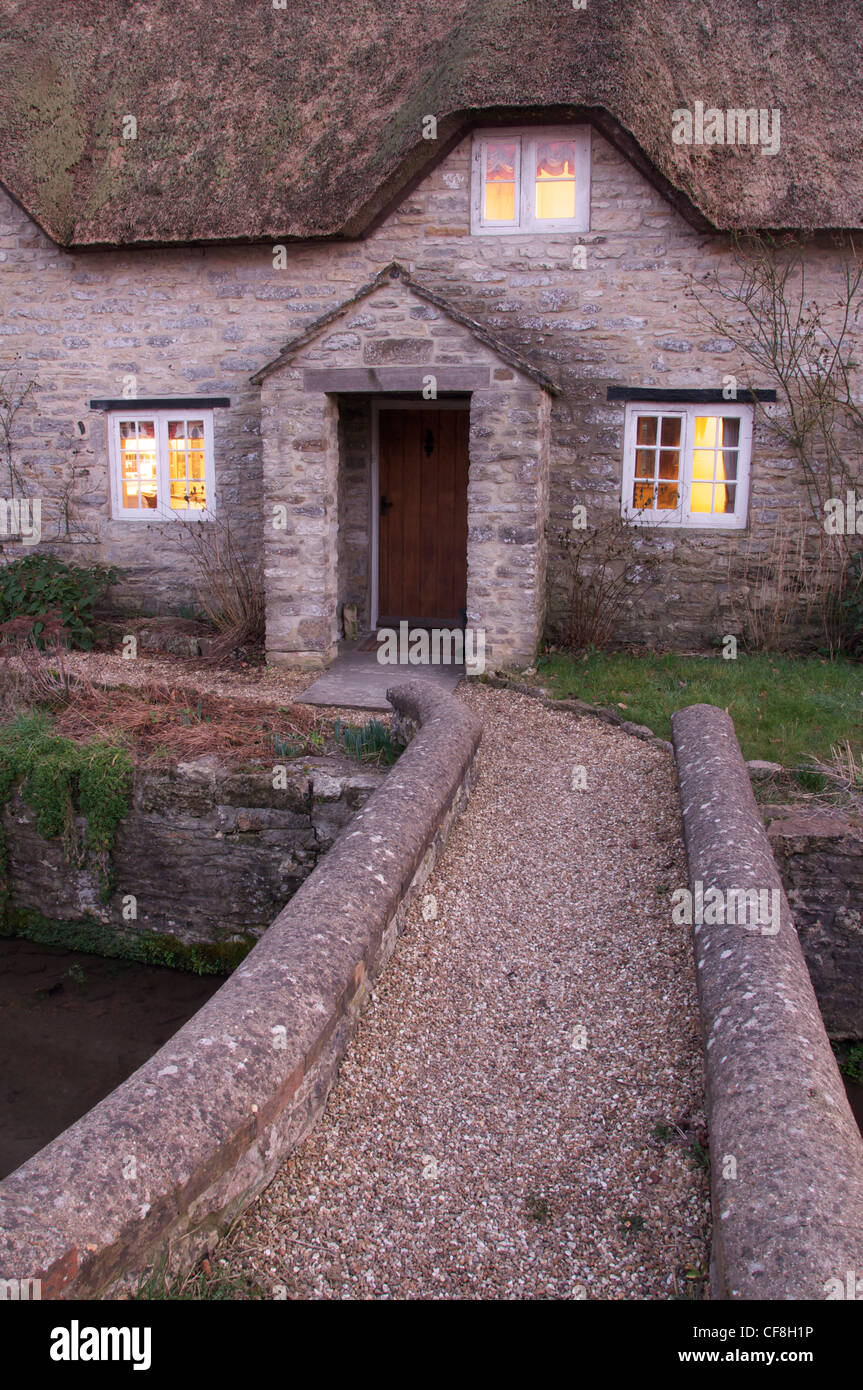 Le luci sono accese in questa pietra pittoreschi cottage con tetto in paglia. Dare un caldo confortante glow in una fredda sera d'inverno. Il Dorset, Inghilterra, Regno Unito. Foto Stock