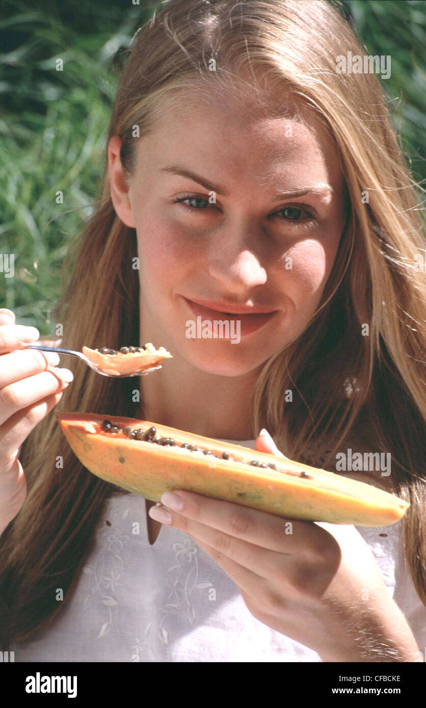 Femmina con lunghi capelli biondi indossa top bianco con broderie anglaise dettaglio, mangiando papaia con cucchiaio cercando di fotocamera a sorridere Foto Stock