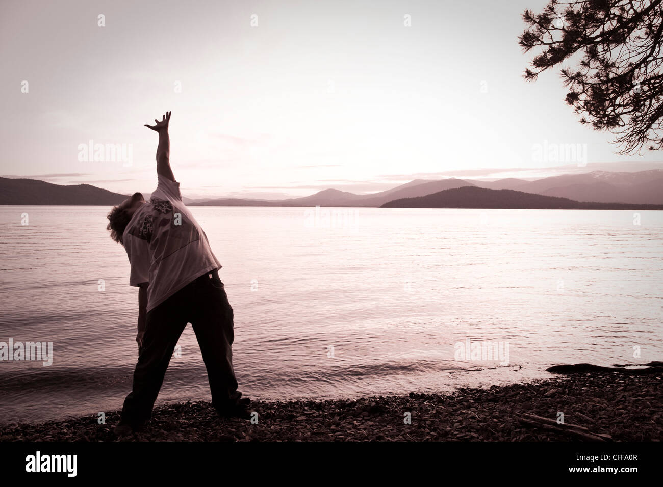 Un uomo archi la schiena con la mano tesa verso il cielo al tramonto accanto a un lago in Idaho. Foto Stock