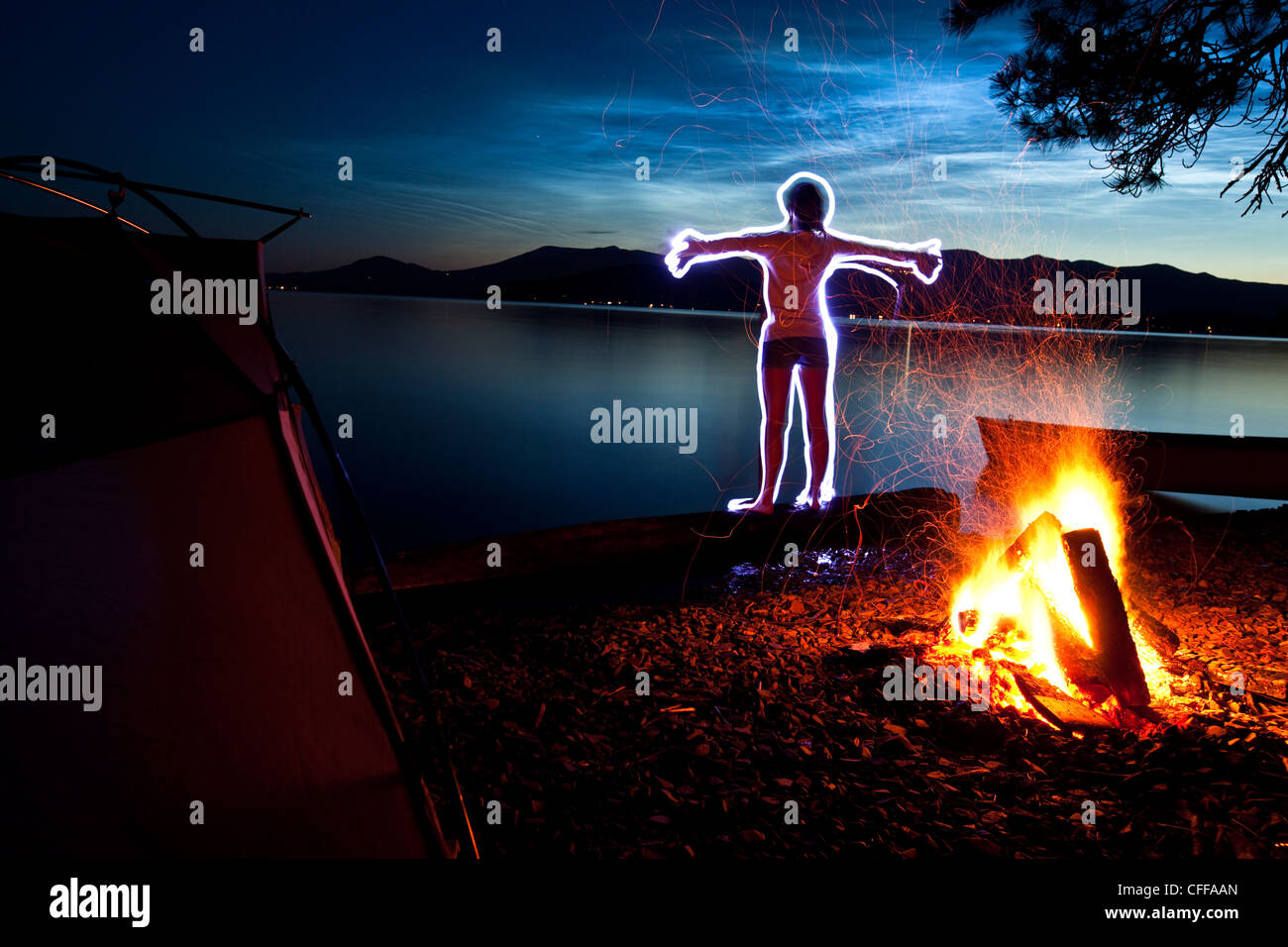 Una pittura di luce di una donna fare yoga accanto alla sua tenda e fuoco su un viaggio di campeggio lungo le rive di un lago in Idaho. Foto Stock