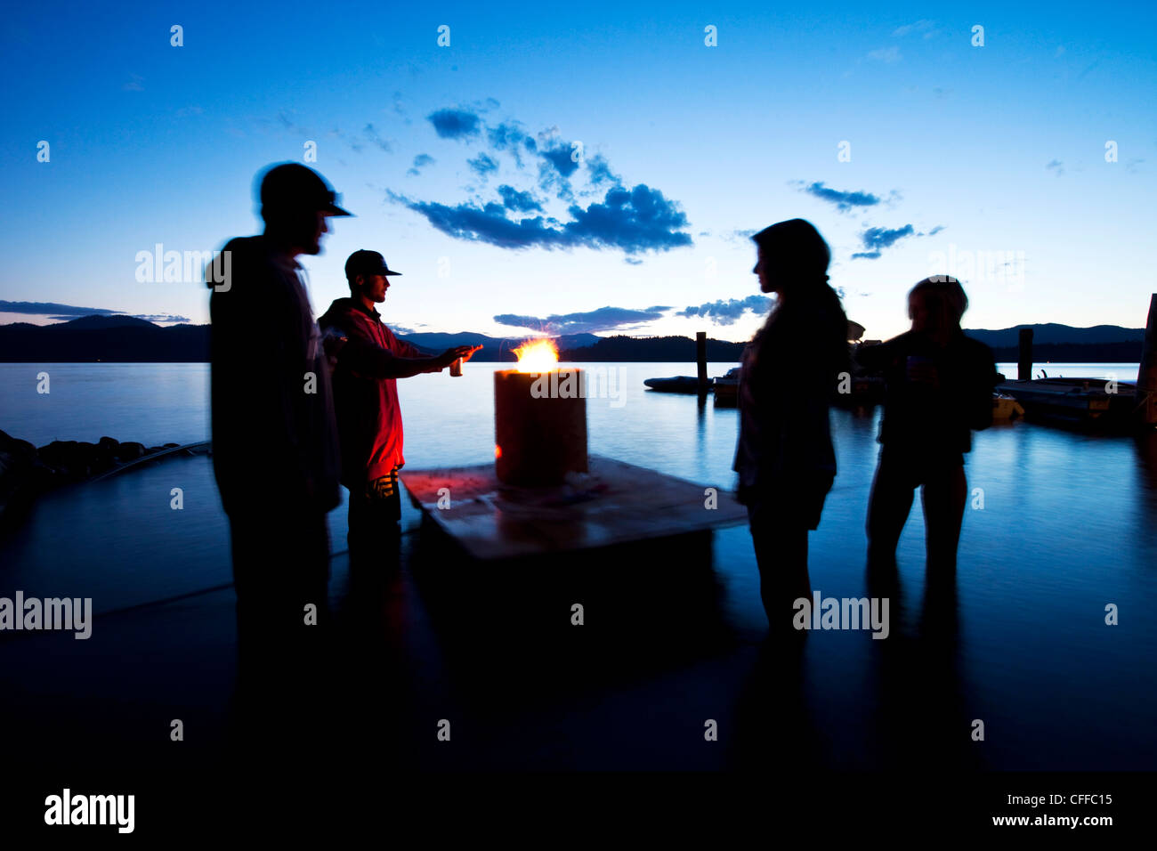 Un gruppo di quattro giovani adulti stand attorno a un fuoco flottante al tramonto in un lago in Idaho. Foto Stock