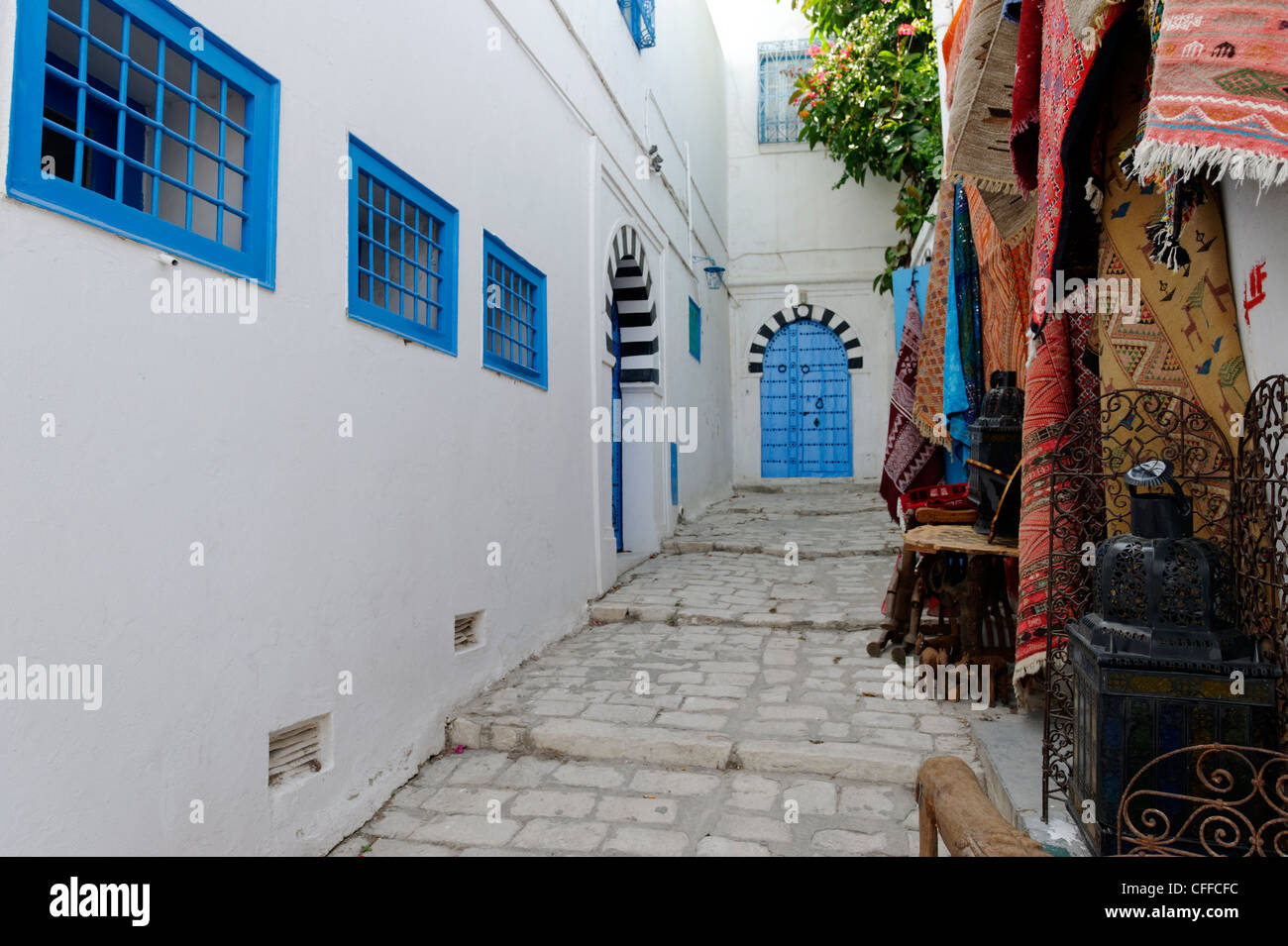 Sidi Bou Said. La Tunisia. Vista dei tappeti di souvenir in vendita lungo stretti vicoli in ciottoli di bianco e nero con blu arcuata in corrispondenza della porta Foto Stock