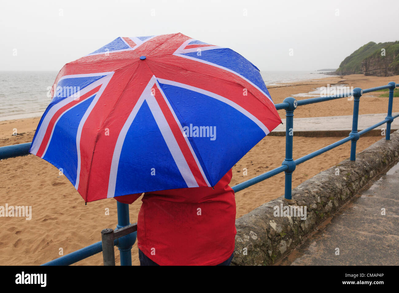 Una donna si affaccia al mare sul lungomare di mare deserta con una Unione Jack ombrello durante molto bagnato estate britannica meteo. Wales UK Foto Stock