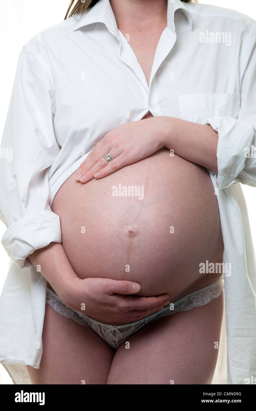 Una giovane donna incinta mentre tiene il suo pancino indossa pantaloni bianchi e un mans camicia bianca su sfondo bianco Foto Stock
