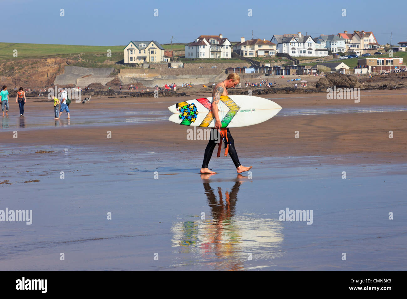 Il surfer che porta una tavola da surf in tutta Crooklets Beach con riflessi nella sabbia bagnata in Bude, North Cornwall, Inghilterra, Regno Unito, Gran Bretagna Foto Stock
