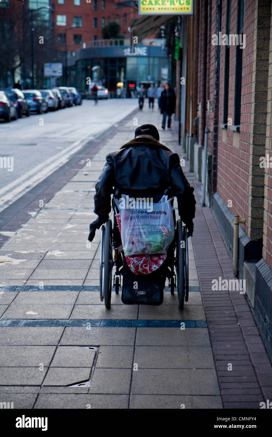 Uomo in sedia a rotelle con le borse della spesa sul retro lottando fino ad un sentiero Sheffield South Yorkshire Inghilterra Foto Stock