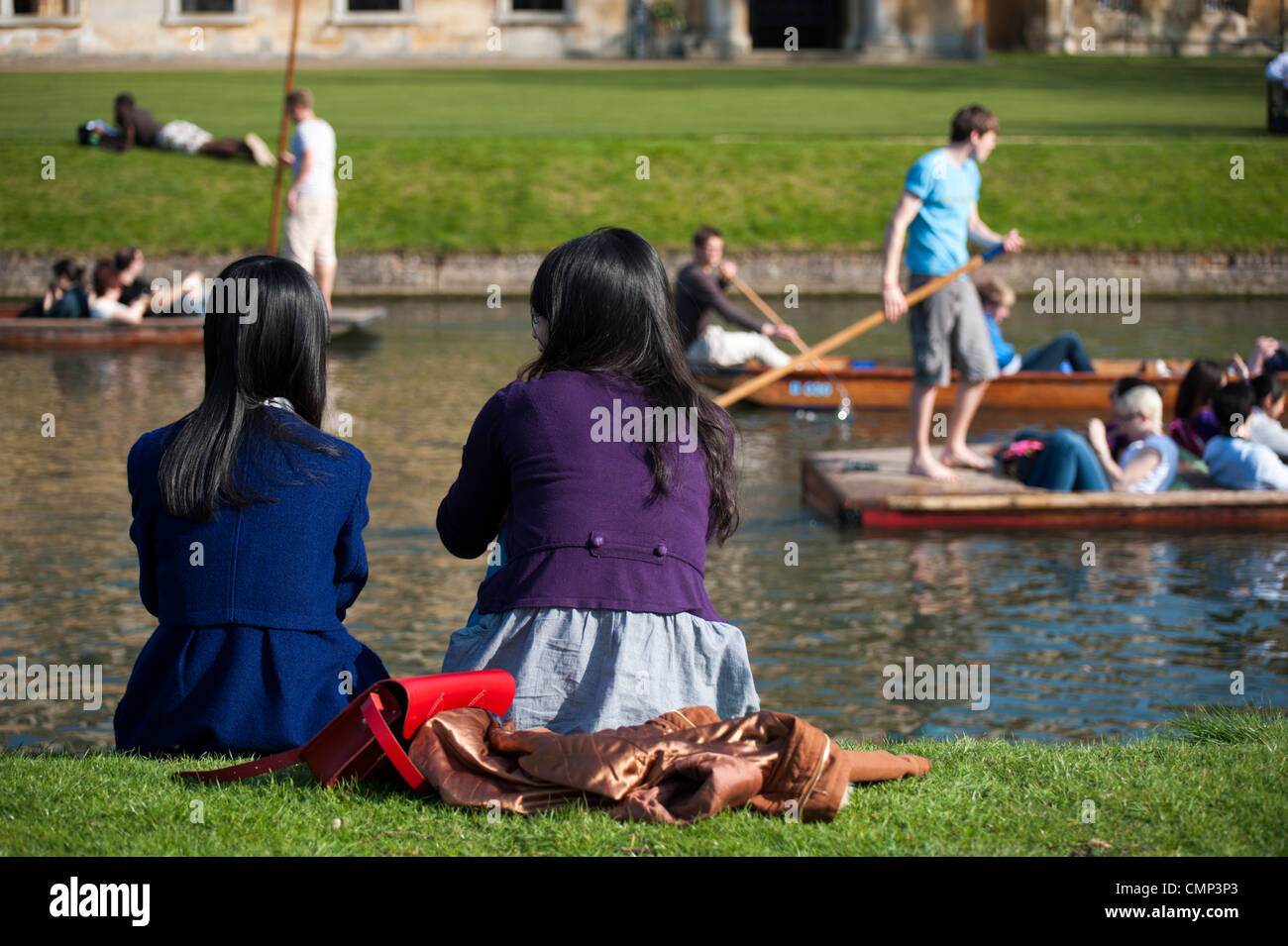 Vista posteriore di due ragazze seduti sulla riva del fiume Cam Cambridge con punting persone in background. Foto Stock