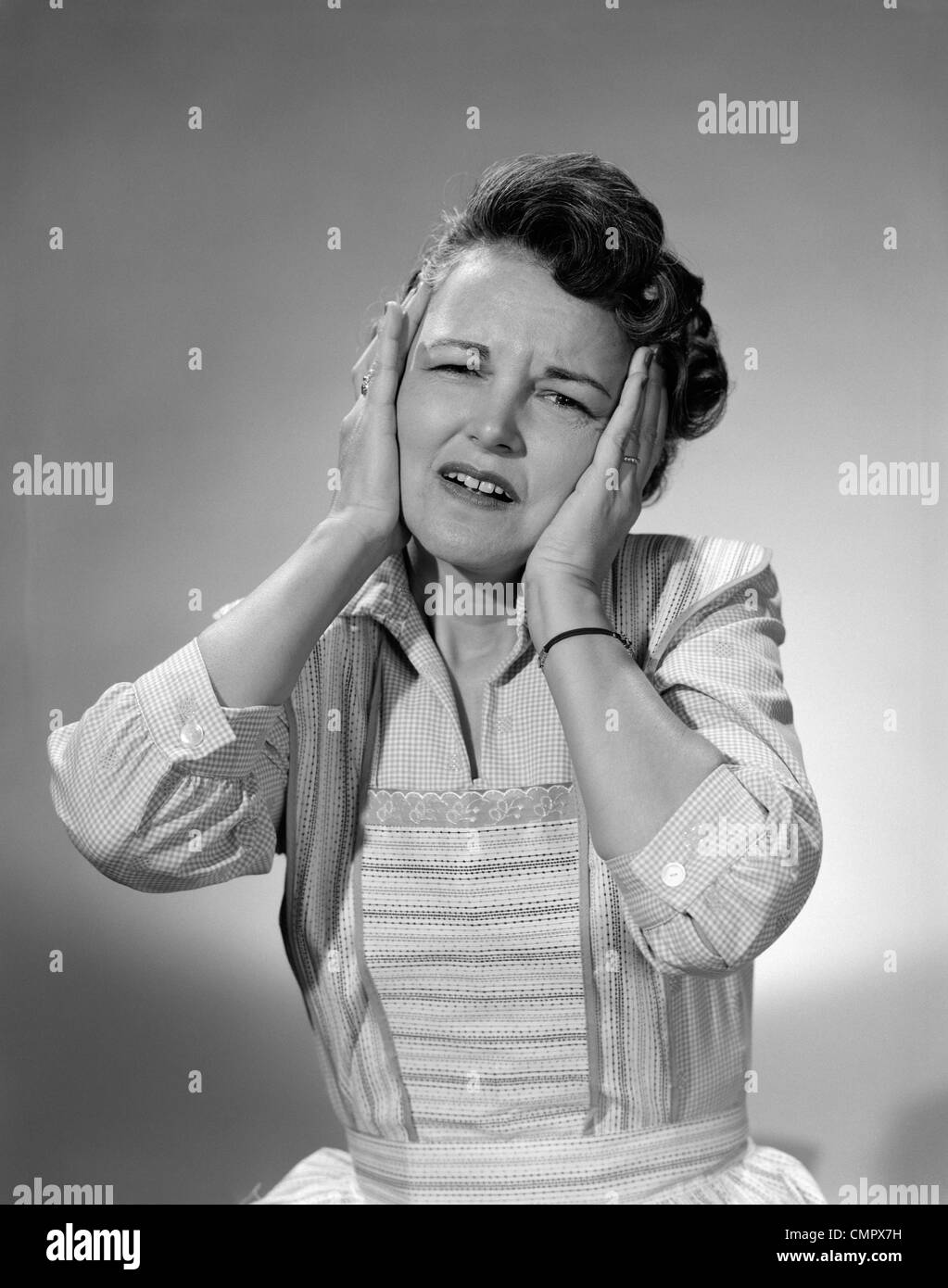 Anni Cinquanta donna in cotone abito con entrambe le mani sopra le sue orecchie tenendo la sua testa accigliata Foto Stock