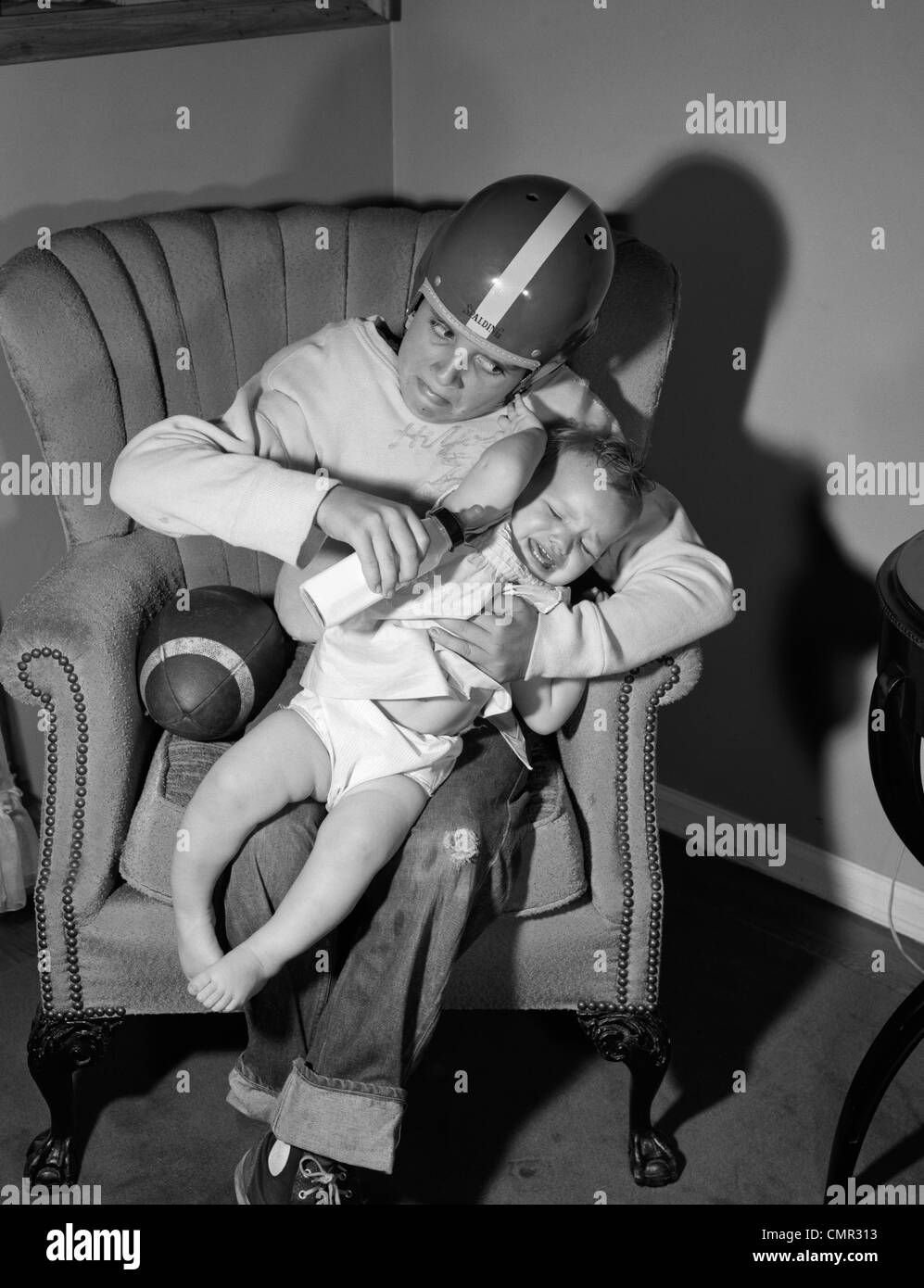 Anni Cinquanta anni sessanta ragazzo seduto da calcio in poltrona indossando il casco cercando di tenere e bottiglia di alimentazione per Gridando bambino Foto Stock