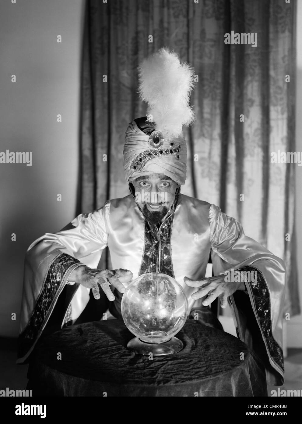 Anni Cinquanta ritratto uomo indovino SWAMI FORTUNE TELLER indossare accappatoio E PIUMA IN turbante guardando sopra la sfera di cristallo Foto Stock