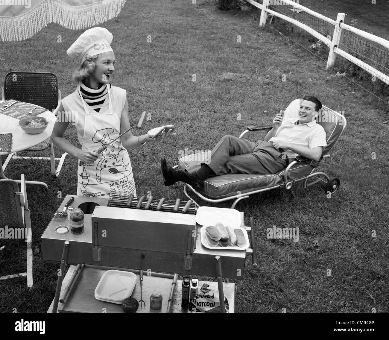 Anni Cinquanta anni sessanta la donna a chef del cappello e grembiule grigliare hot dogs sul grill UOMO IN CHAISE LONGUE a ridere di mangiare un FRANKFURTER AL DI FUORI Foto Stock