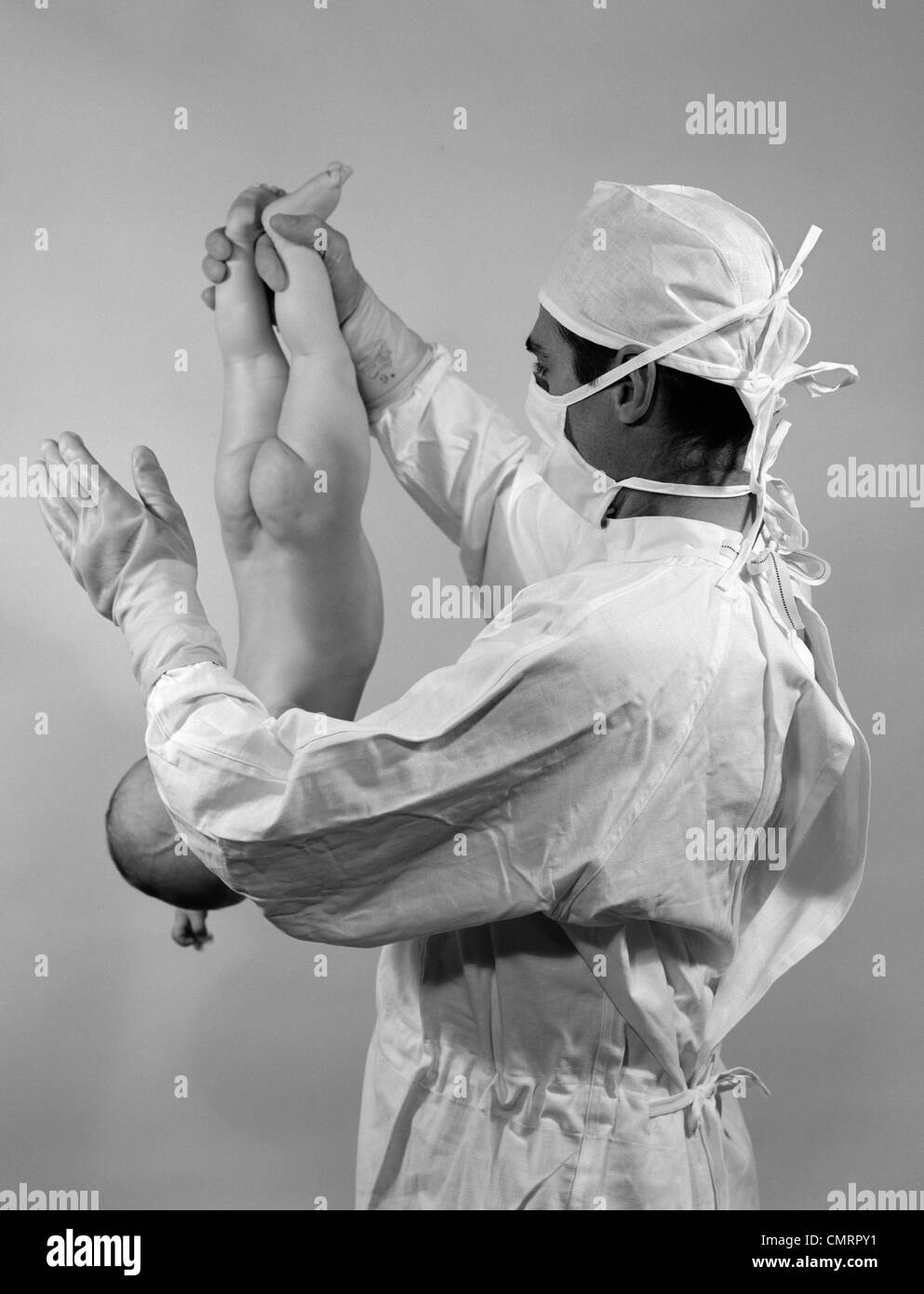 Anni sessanta uomo medico tenendo neonato capovolto mediante caviglie SLAPPING FONDO PER AVVIARE lo stereotipo di respirazione del processo di nascita Foto Stock