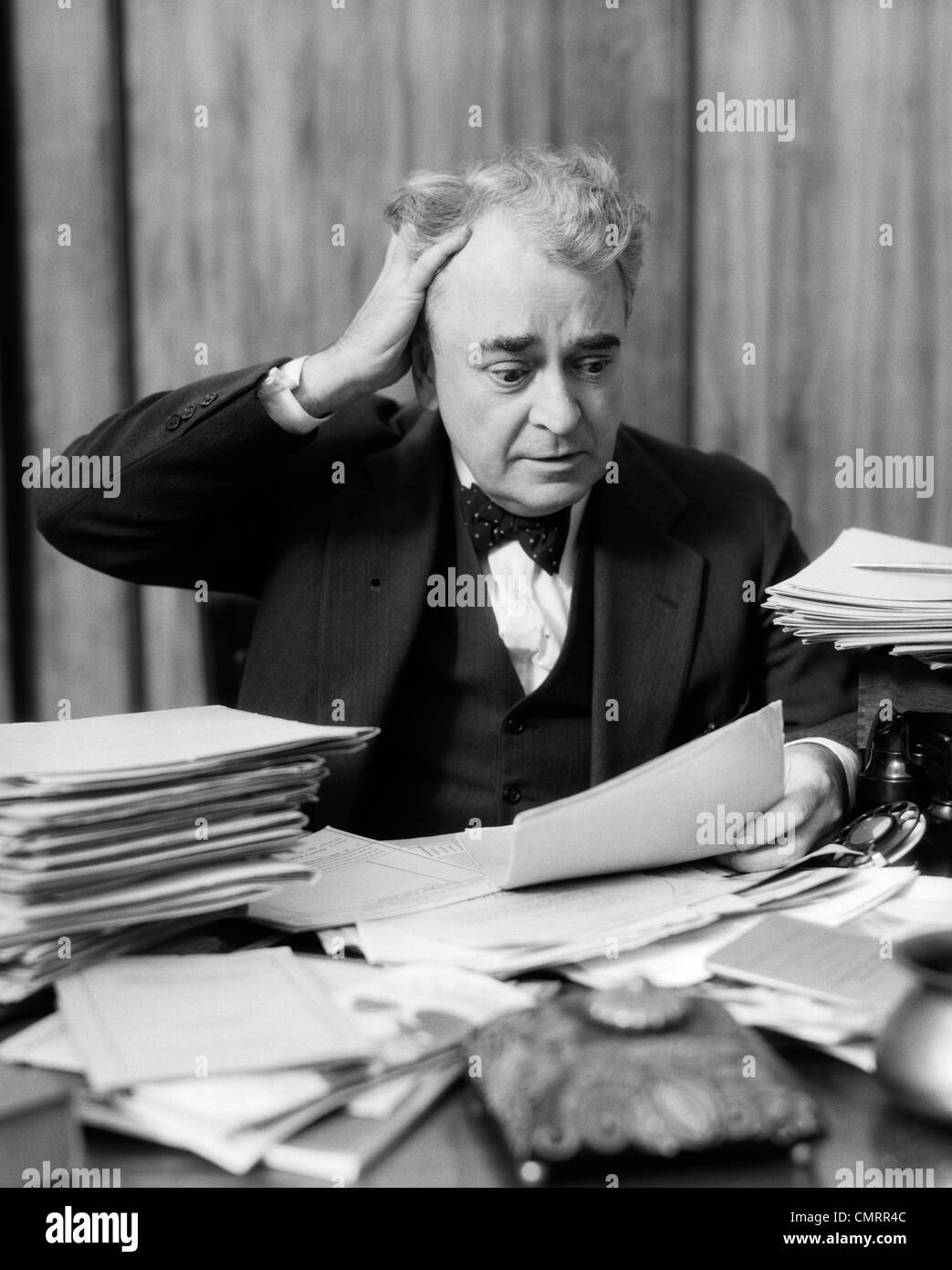 1930s imprenditore anziani seduti alla scrivania tra pile di carta con le mani nei capelli & espressione sconcertata Foto Stock