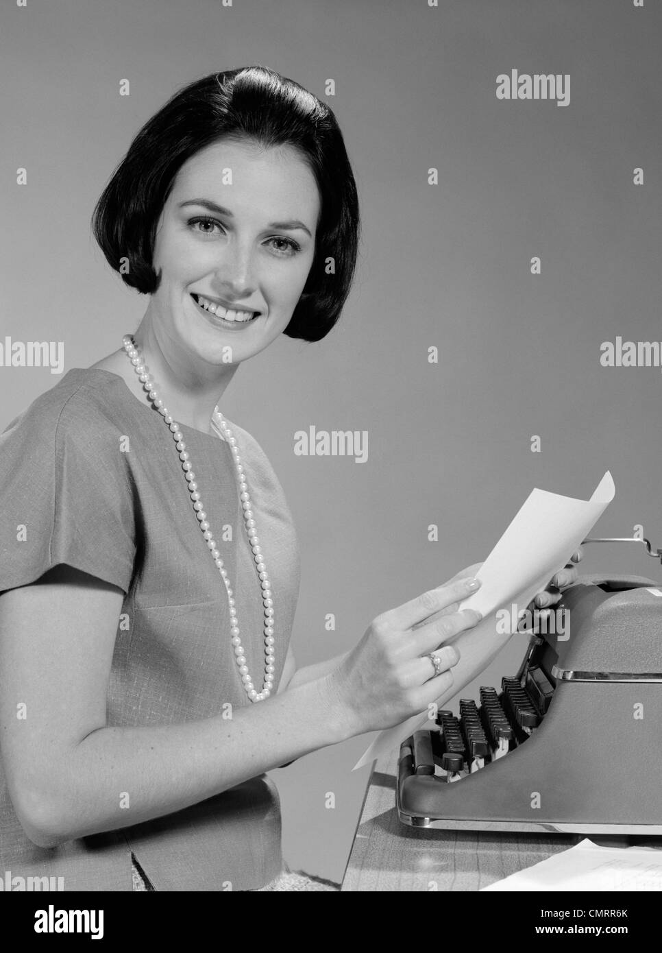 Anni sessanta donna Segretario sorridente seduto alla scrivania con manuale dattiloscritto guardando la fotocamera Foto Stock