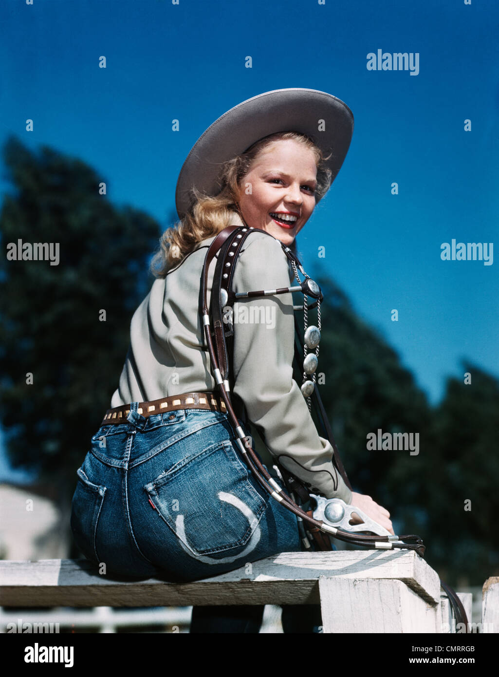 Negli anni quaranta anni cinquanta sorridente donna bionda che indossa WESTERN COWGIRL vestiti guardando sopra la spalla seduto sulla recinzione di legno Foto Stock