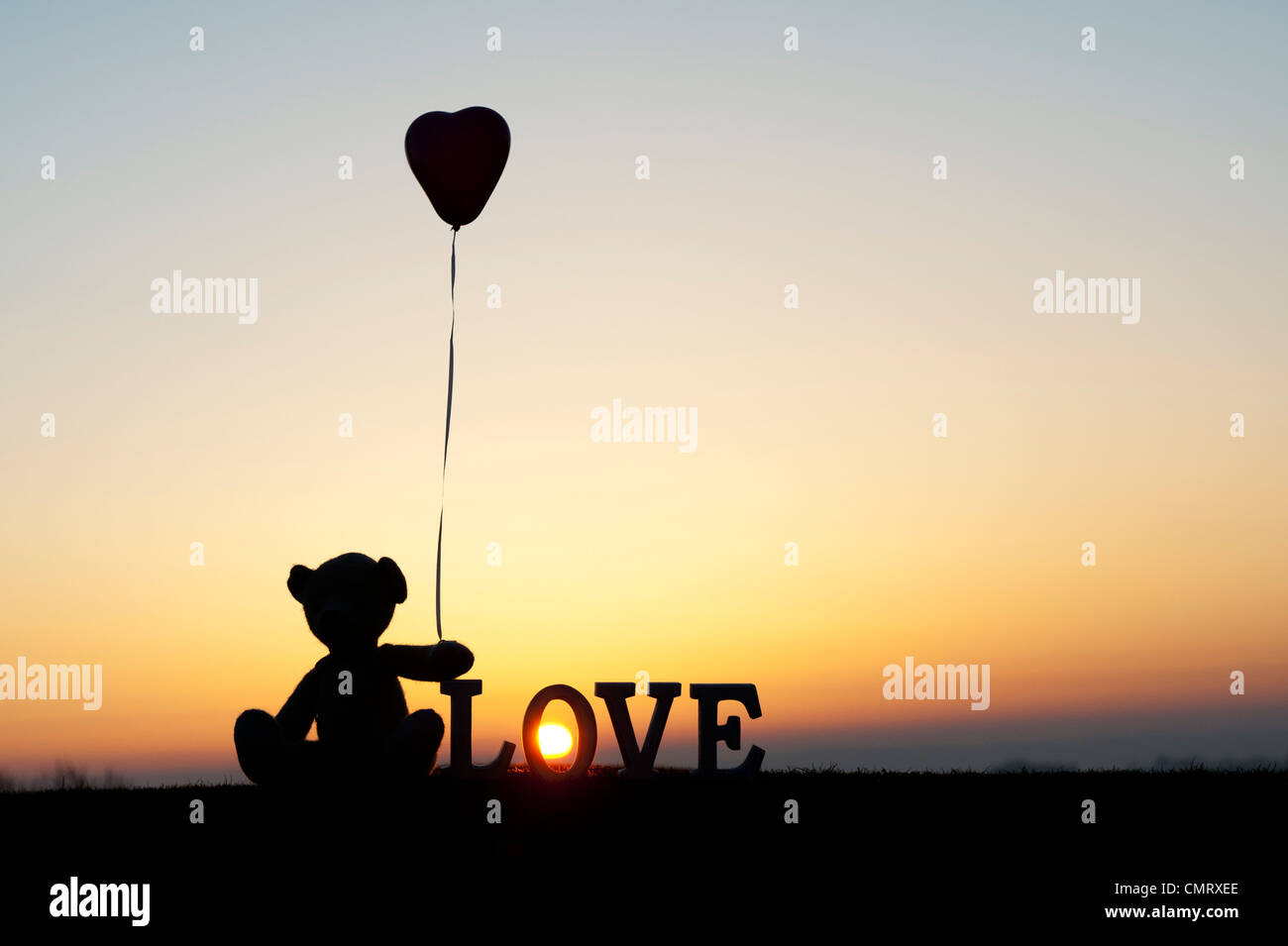 Orsacchiotto di peluche tenendo a forma di cuore pallone avanti anche la parola amore a sunrise Foto Stock