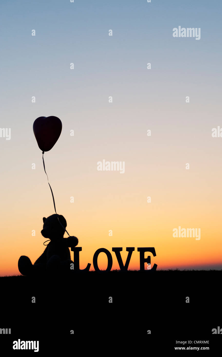 Orsacchiotto di peluche tenendo a forma di cuore pallone avanti anche la parola amore a sunrise Foto Stock