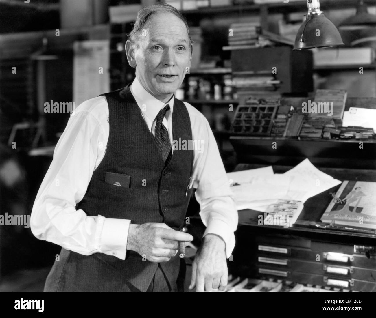 1930s 1940s vecchio uomo che indossa SHIRTSLEEVES VEST appoggiata sul cassetto di tipo armadio nel vecchio negozio di stampa di fumare un buon sigaro e parlare Foto Stock