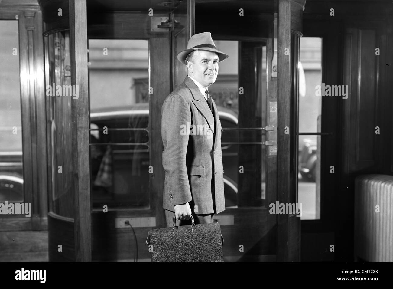 1930s 1940 imprenditore sorridente IN HAT doppio petto tuta valigetta lasciando edificio attraverso la porta girevole guardando la fotocamera Foto Stock