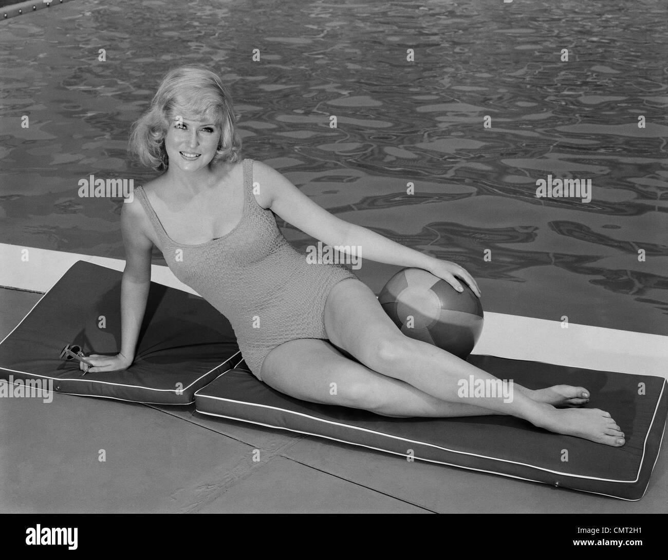 Anni sessanta sorridente DONNA BIONDA indossando il costume da bagno in piscina che pongono la mano sulla palla spiaggia Foto Stock