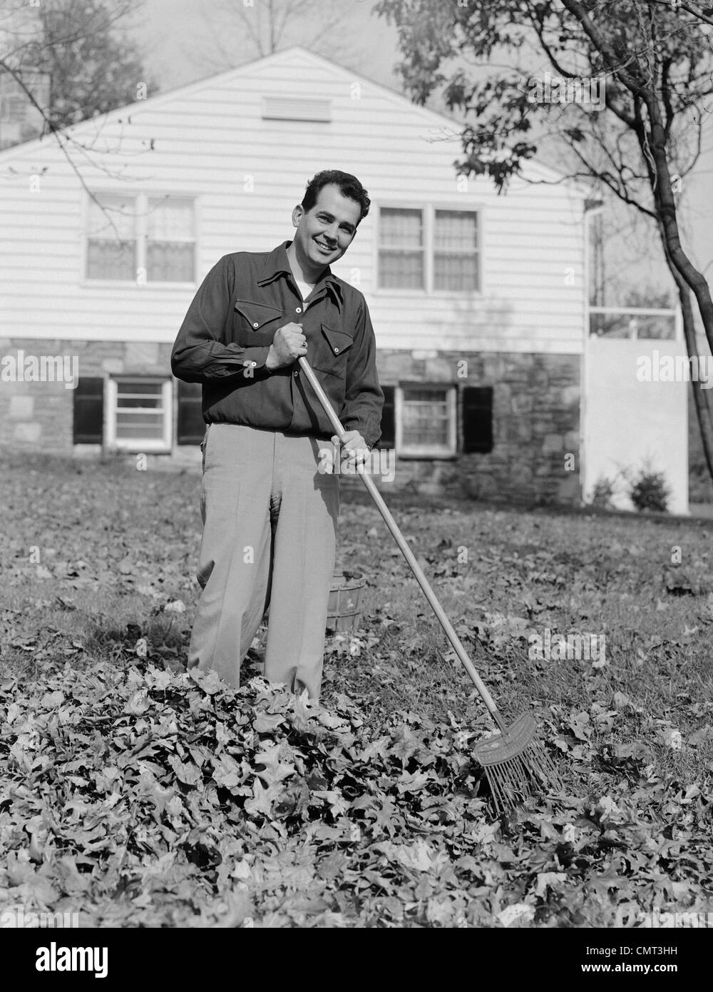 Anni Cinquanta anni sessanta uomo sorridente a rastrellare foglie di autunno nel cortile anteriore della casa Foto Stock