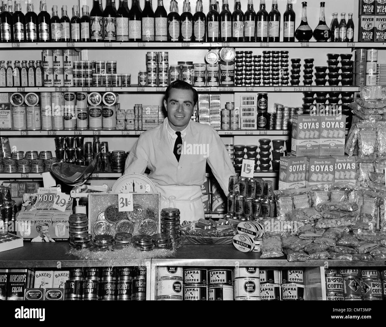 1940s uomo sorridente drogheria in piedi dietro il contatore RIEMPITO CON VARI PRODOTTI ALIMENTARI IN Contenitori e imballaggi interni negozio di alimentari Foto Stock