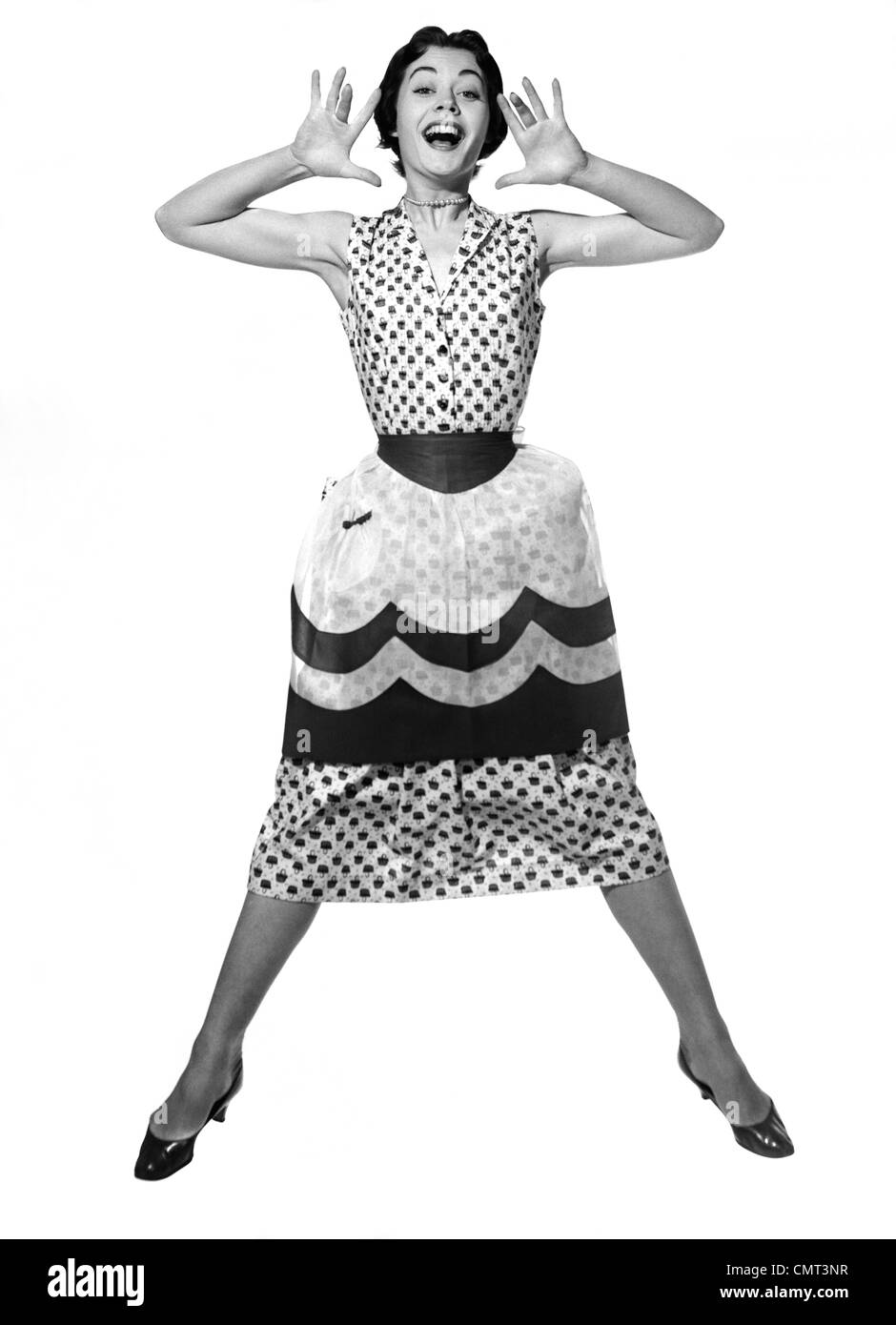 Anni Cinquanta LA DONNA IN ABITO & grembiule salti in aria gambe divaricate & Palme diffusione su entrambi i lati della faccia con la bocca aperta guardando la fotocamera Foto Stock