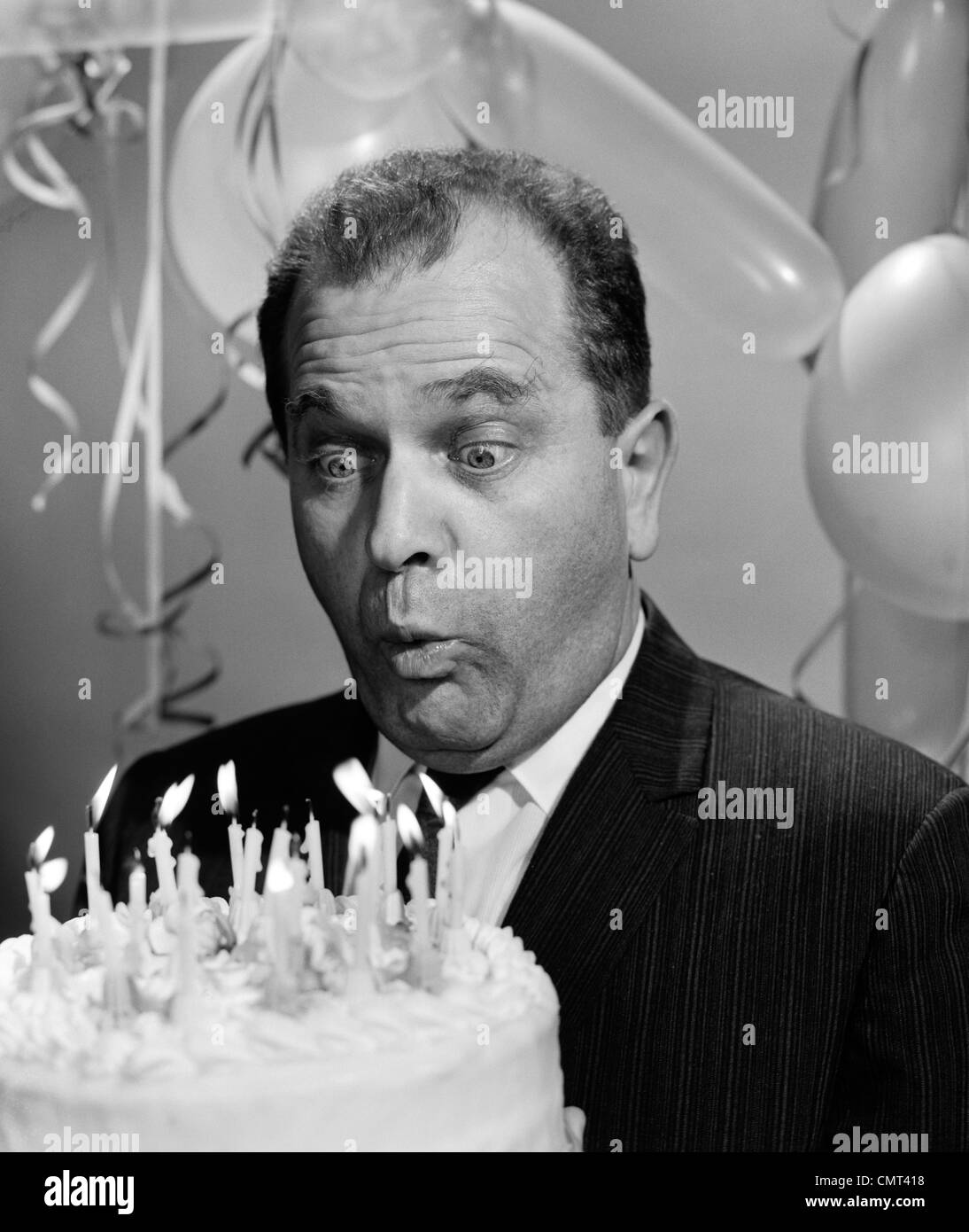 Anni sessanta uomo soffiando le candeline sulla torta di compleanno con occhi sporgenti e palloncini IN BACKGROUND Foto Stock