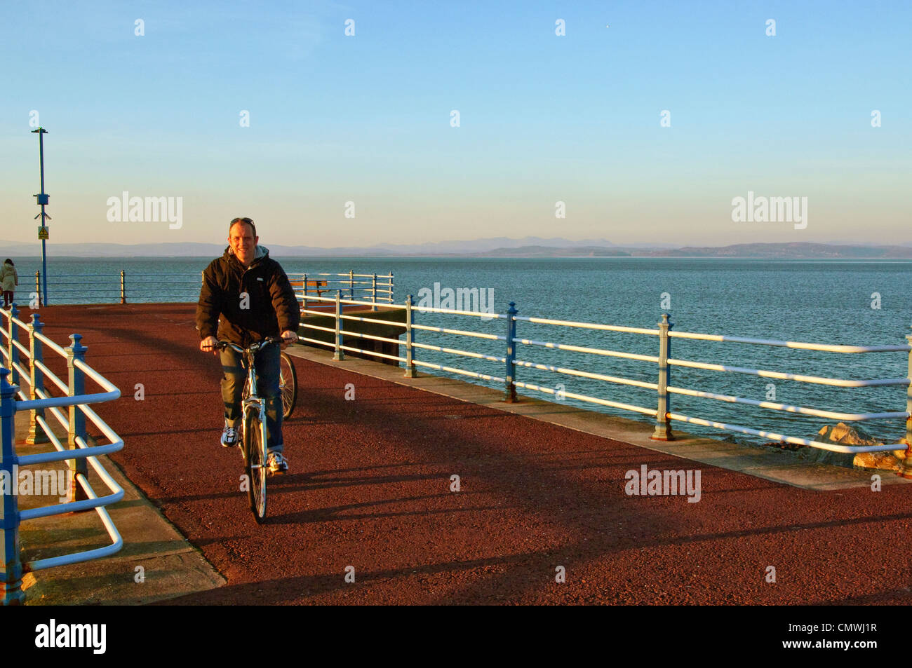 Ciclista sul molo di pietra, Morecambe, Lancashire, con Morecambe Bay e Lakeland Fells oltre su una sera d'inverno Foto Stock