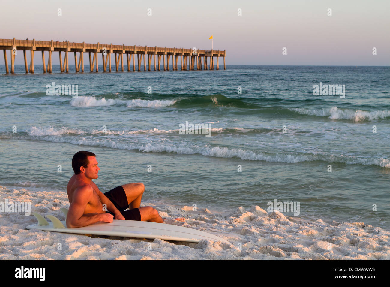 I giovani adulti surfer siede sulla spiaggia appoggiata sulla sua tavola da surf con un molo in background sulla spiaggia di Pensacola, Florida. Foto Stock