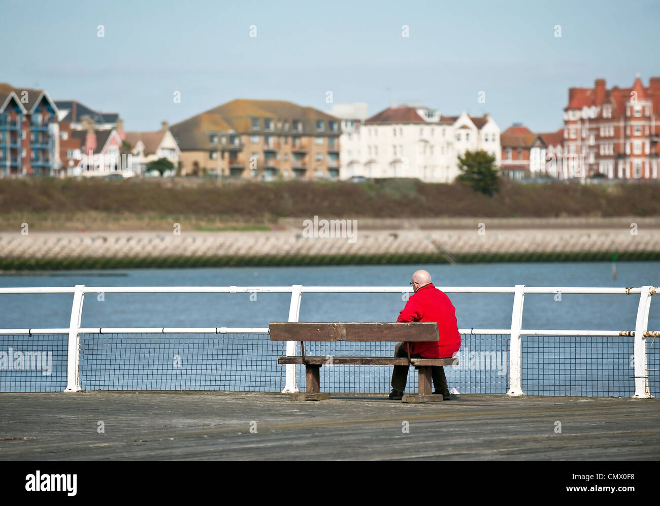 Un uomo con una giacca rossa seduto su una panchina sul molo di Clacton sulla costa dell'Essex. Foto Stock
