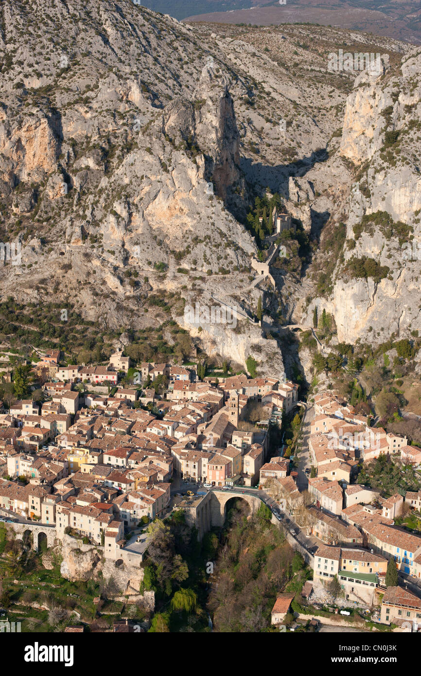 VISTA AEREA. Borgo medievale ai piedi di una massiccia scogliera. Moustiers-Sainte-Marie, Alpes-de-Haute-Provence, Francia. Foto Stock