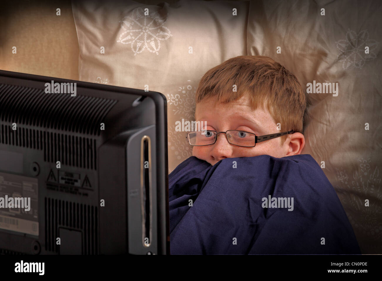 Un ragazzo sembra spaventato come egli guarda la televisione a letto Foto Stock