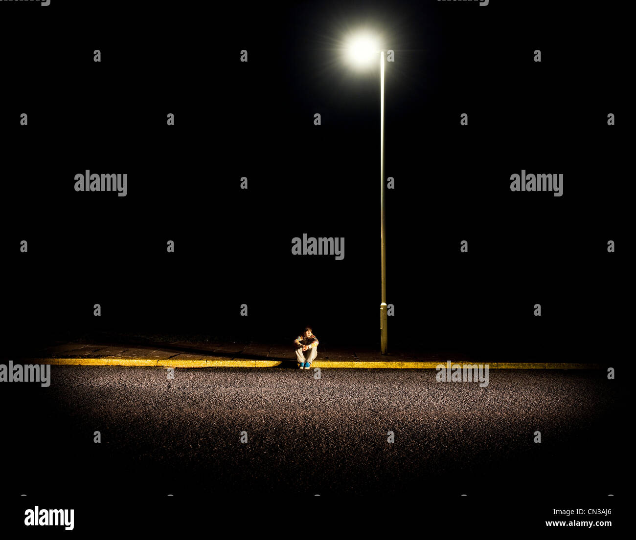 Ragazzo seduto su un marciapiede durante la notte con le luci di strada Foto Stock
