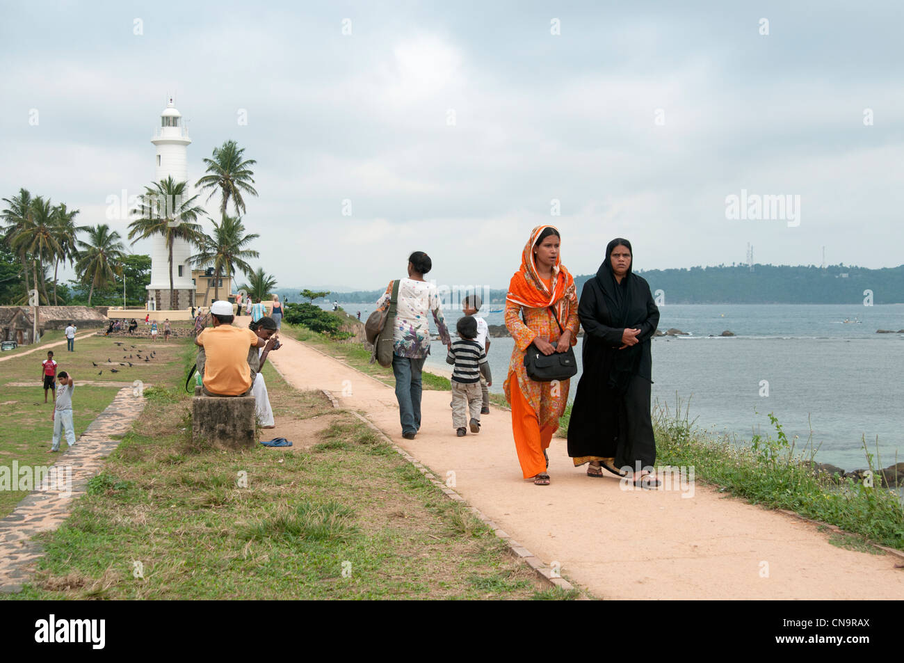 Musulmani e famiglie Indù a piedi lungo le mura del Forte di Galle Sri Lanka con il faro in background Foto Stock